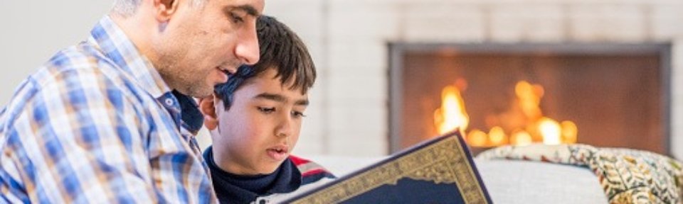 A Muslim father and son are sitting on a couch and reading the Quran together. The fireplace is on in the background.